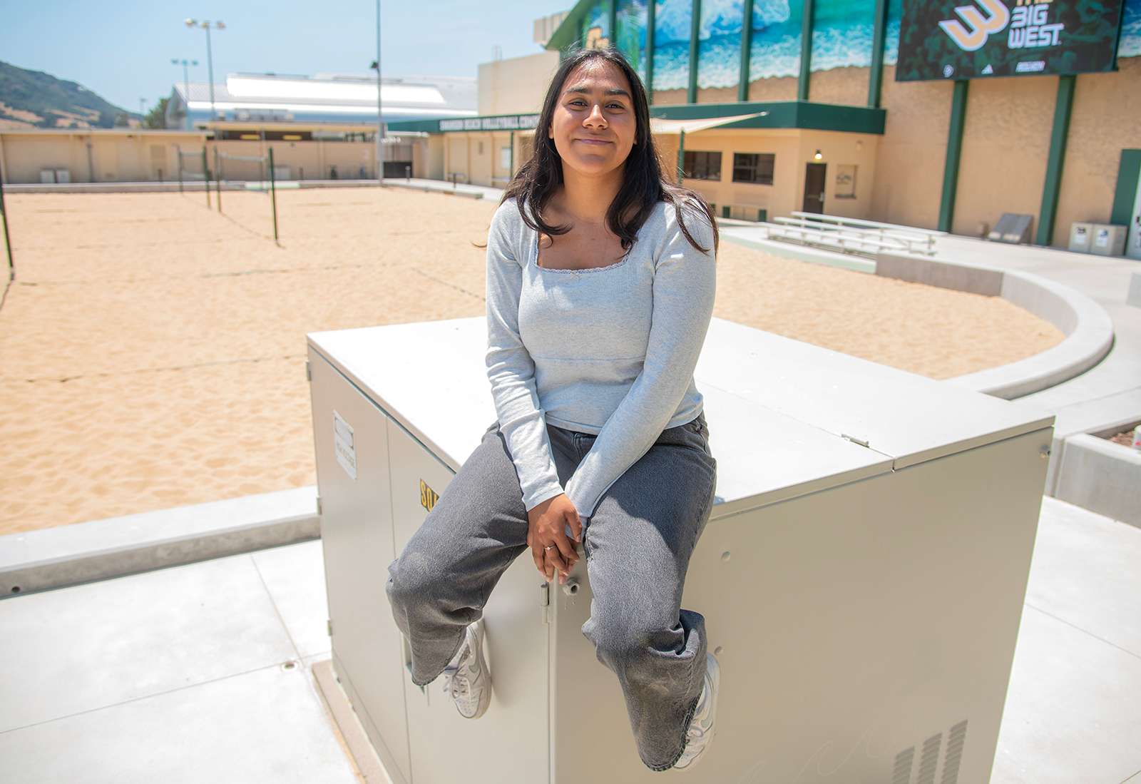Young woman wearing gray long-sleeve shirt and dark gray pants sitting on an outdoor utility box near sand volleyball courts.