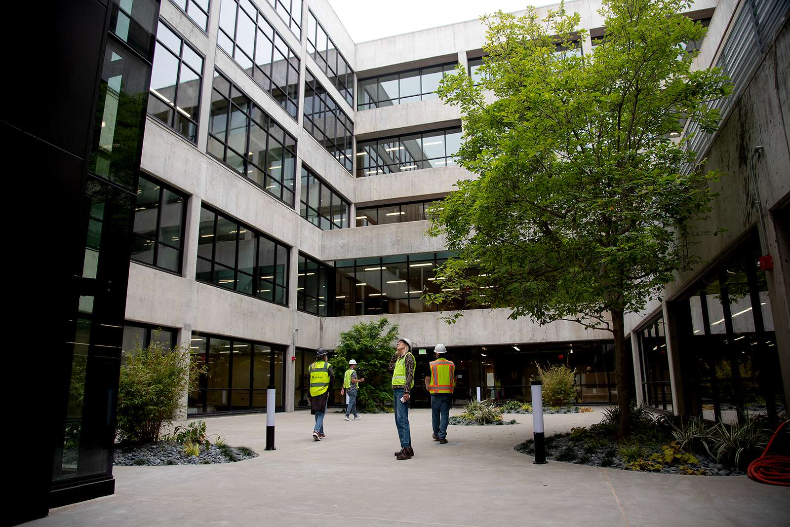 Construction workers wearing safety vests and hard hats walk through a newly renovated courtyard surrounded by multi-story concrete and glass buildings, with landscaping and young trees planted throughout the space.