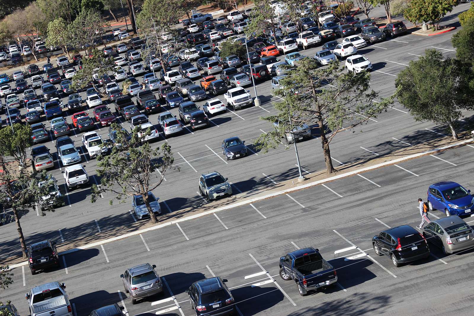 A large parking lot with many parked cars, some empty spaces, trees, and a person walking with an orange backpack.