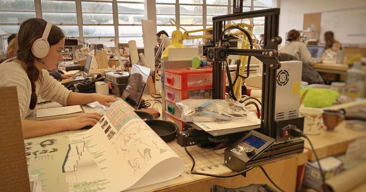 Woman with headphones working on a laptop and architectural drawings beside a 3D printer in a busy office.