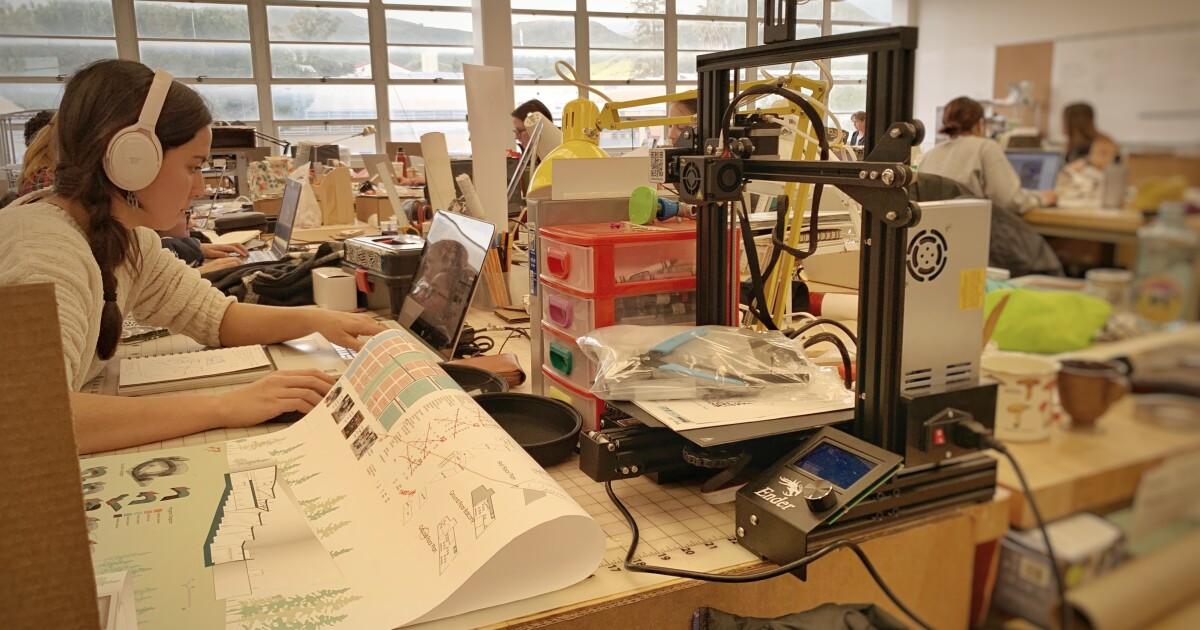 Woman with headphones working on a laptop and architectural drawings beside a 3D printer in a busy office.