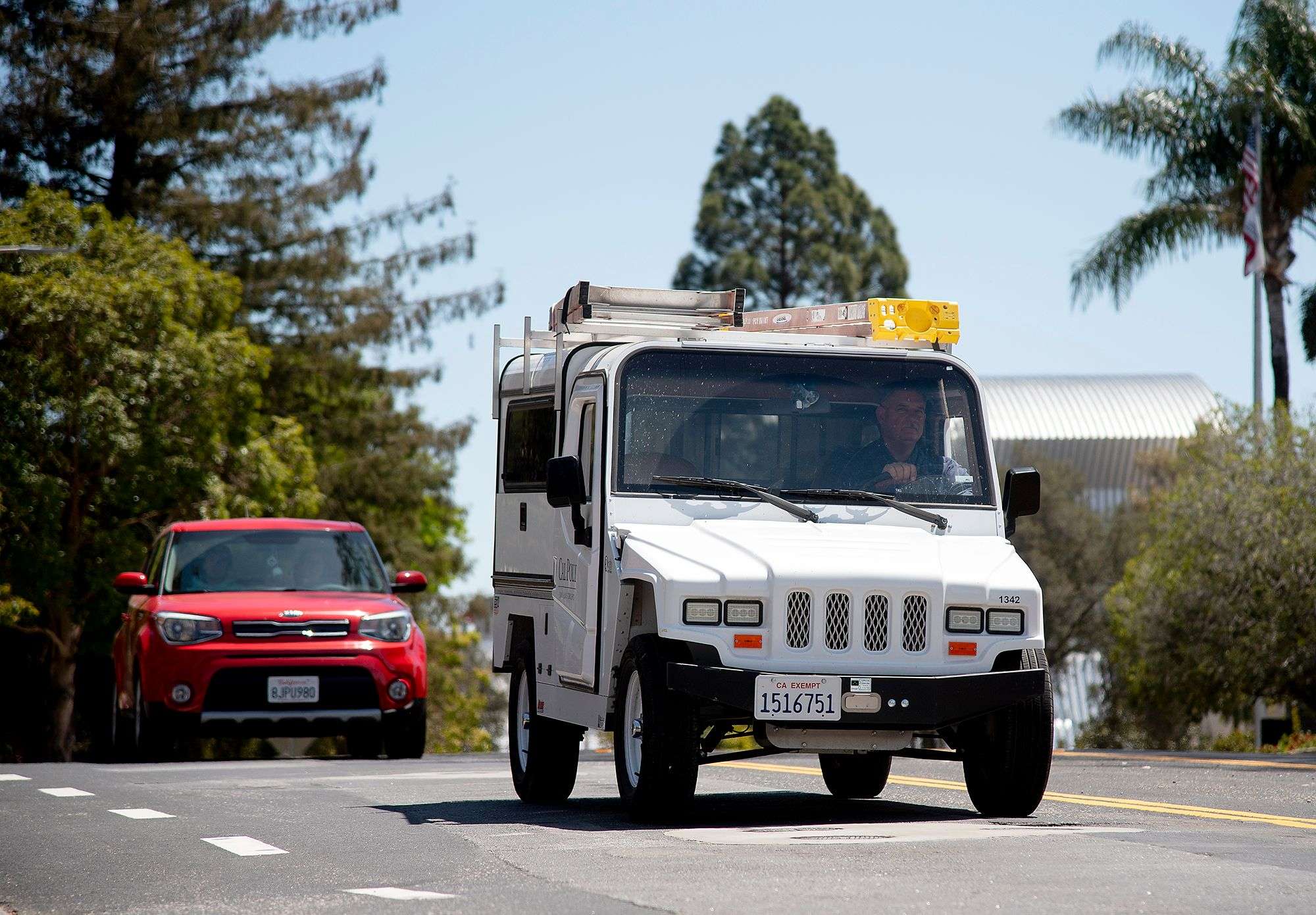 White utility truck with ladders on top driving on a street, followed by a red car, trees and buildings in background.