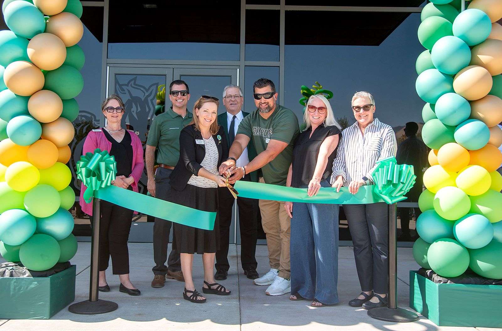 Group of seven people cutting a green ribbon in front of a building entrance decorated with green and yellow balloons.