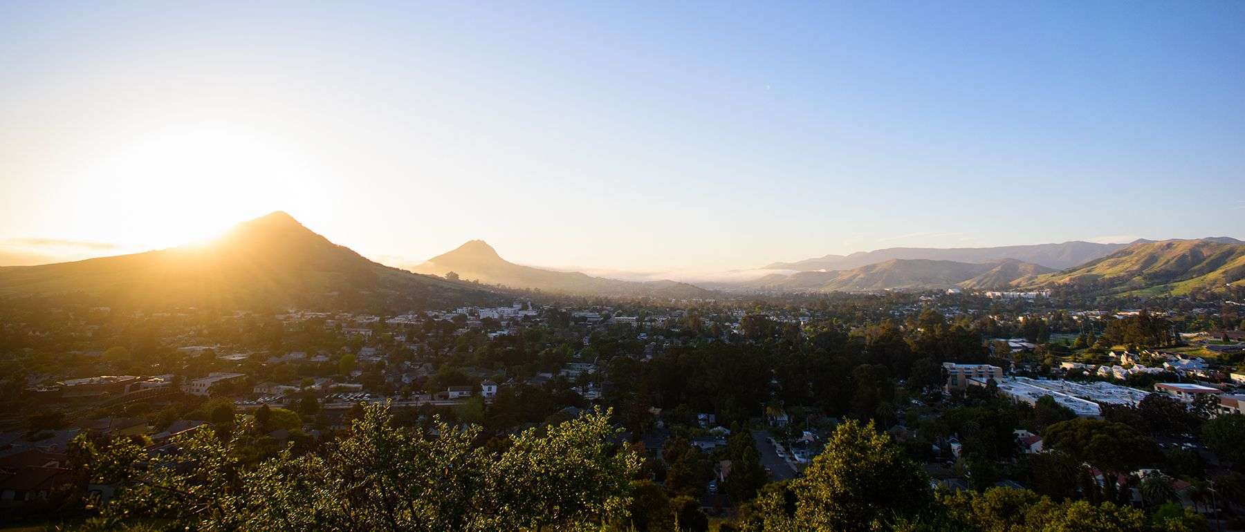 Sun setting behind mountain overlooking a city with scattered trees and buildings.