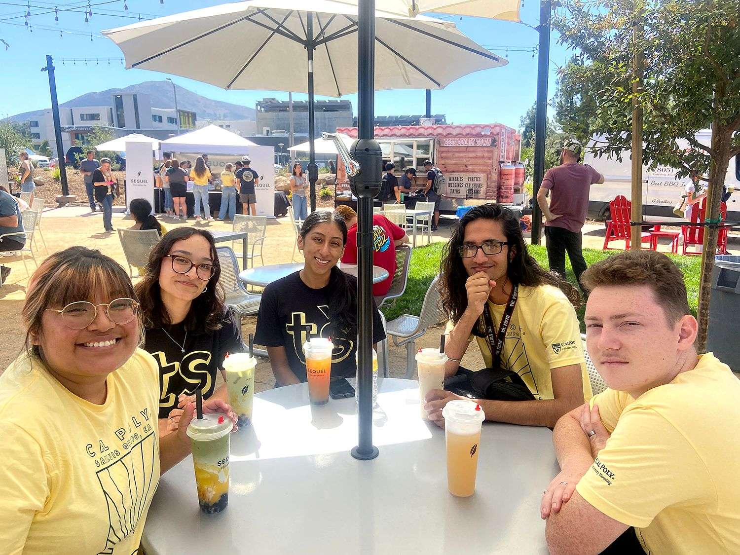 Five people sitting at an outdoor table with drinks under a large umbrella, with food trucks and others in background.