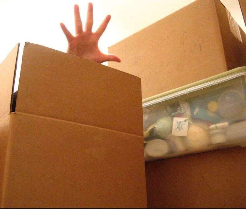 Hand reaching out from behind stacked moving boxes, one transparent box shows various toiletries inside.