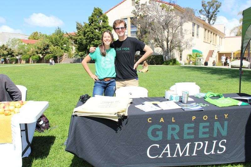 Two people stand behind a table with a Cal Poly Green Campus banner on a sunny lawn.