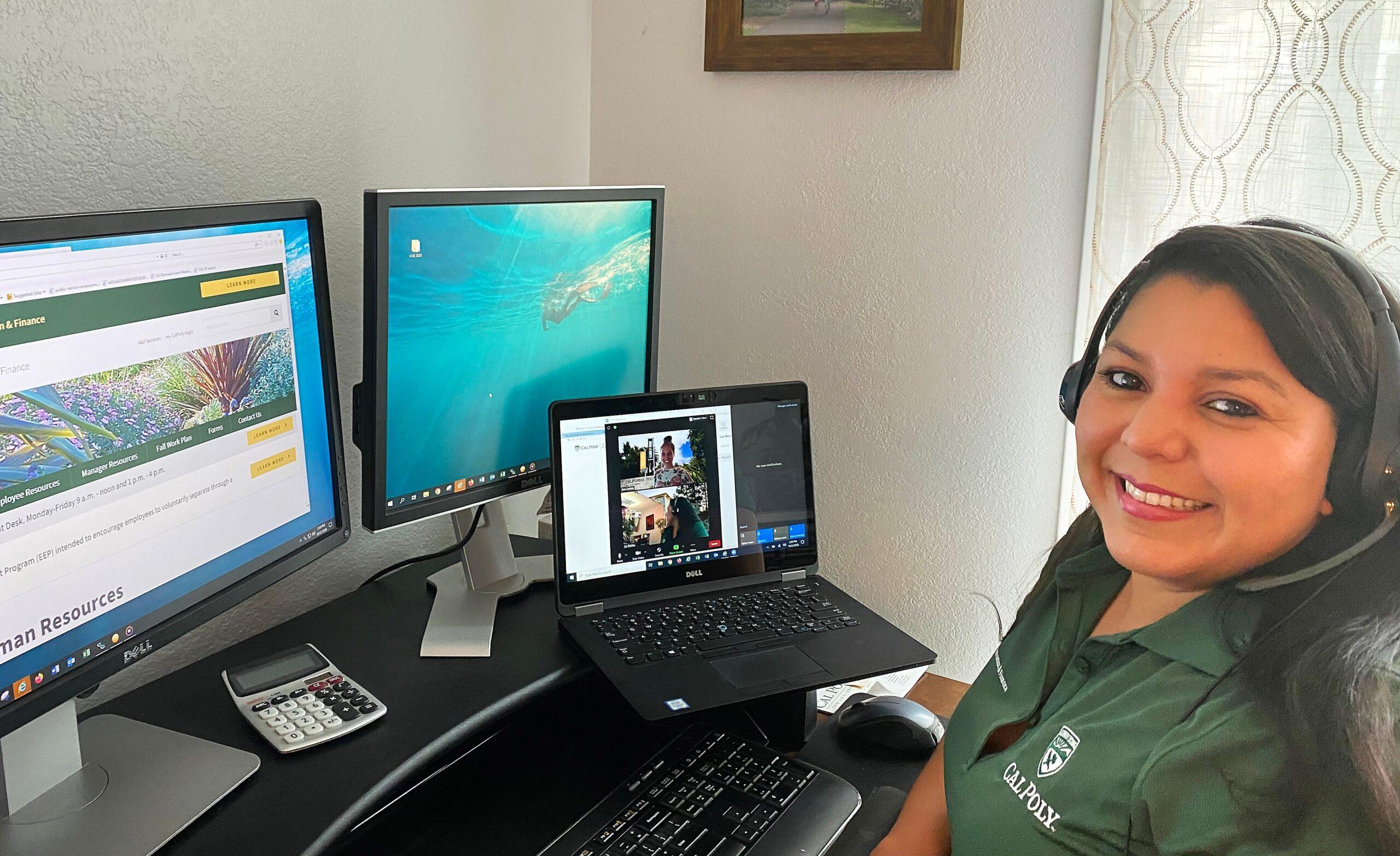 Woman smiling wearing a Cal Poly polo and headset, sitting at a desk with dual monitors and a laptop in a video call.
