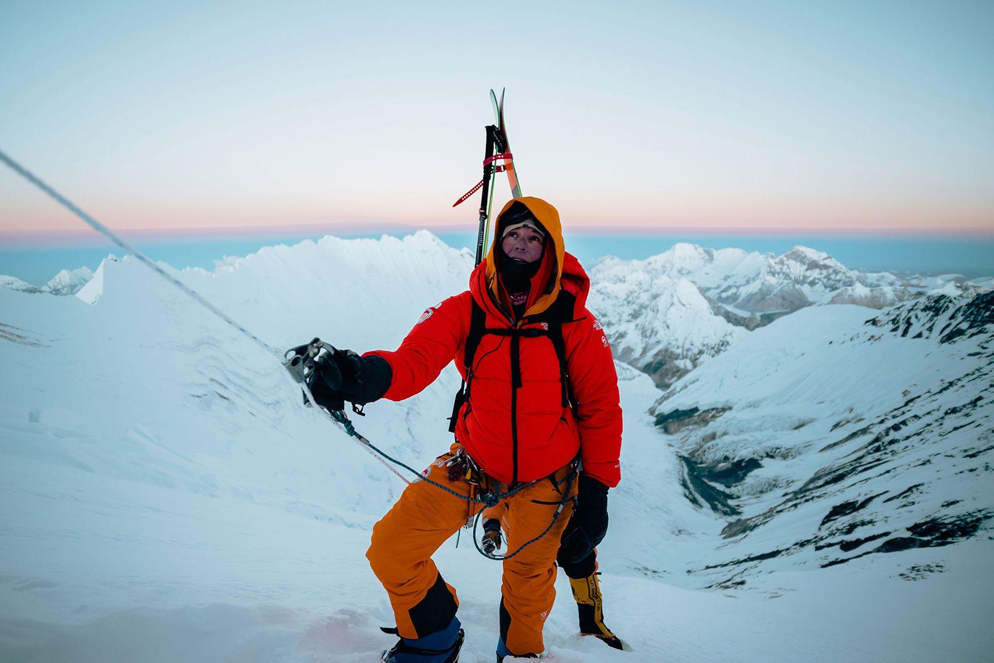 Mountain climber in orange and red gear ascending snowy peak with ski poles and mountain range background at sunrise.