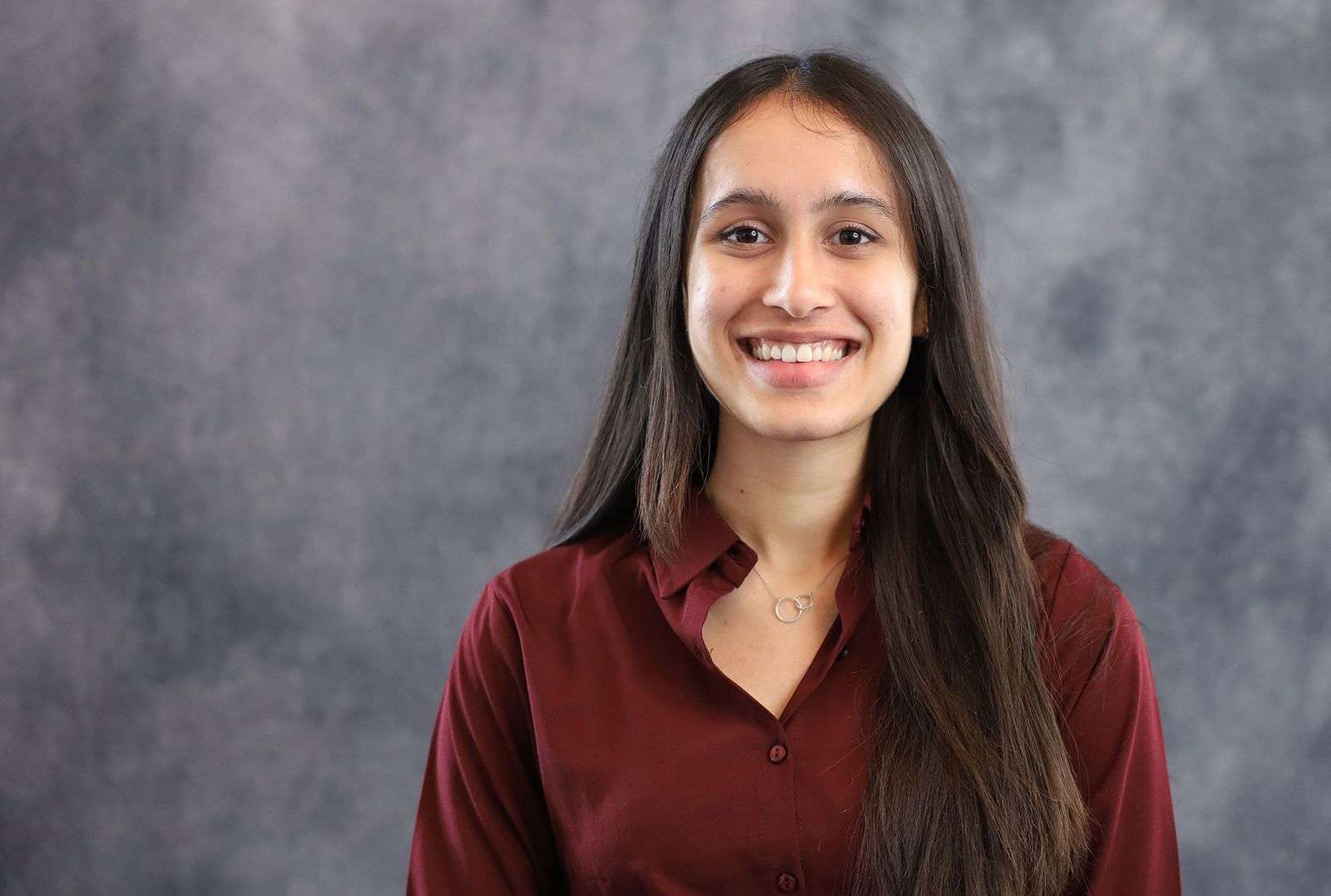 Young woman with long dark hair wearing a maroon blouse, smiling against a gray textured background.