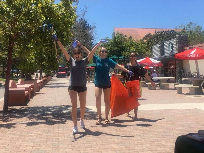 Three women outdoors on a sunny day, holding trash pickers and orange bags, smiling on a paved walkway.