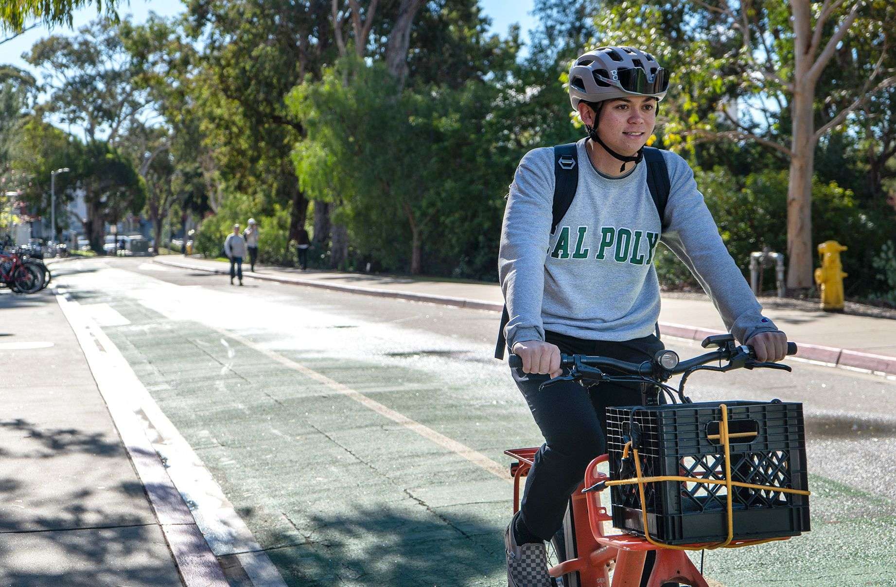 Young man wearing "CAL POLY" sweatshirt and helmet riding a bike with a black crate on a sunny bike lane.