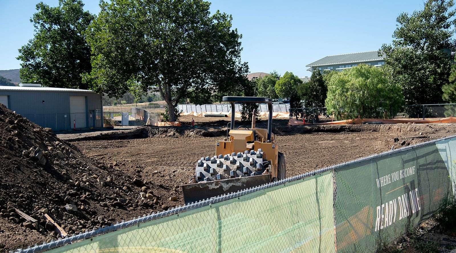 Construction site with soil compactor, dirt piles, fencing, trees, and buildings in the background.