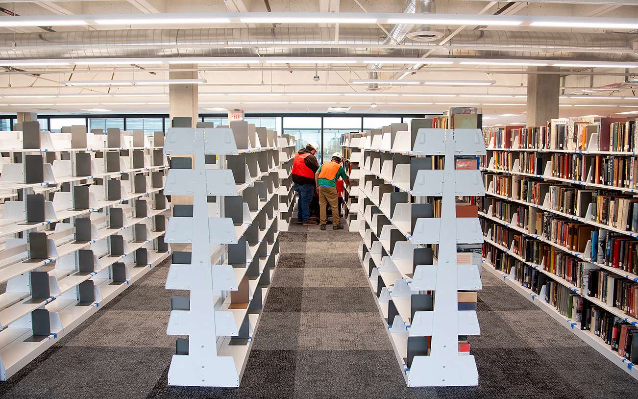 Two people organizing mostly empty white bookshelves in a brightly lit library aisle with one fully stocked shelf.