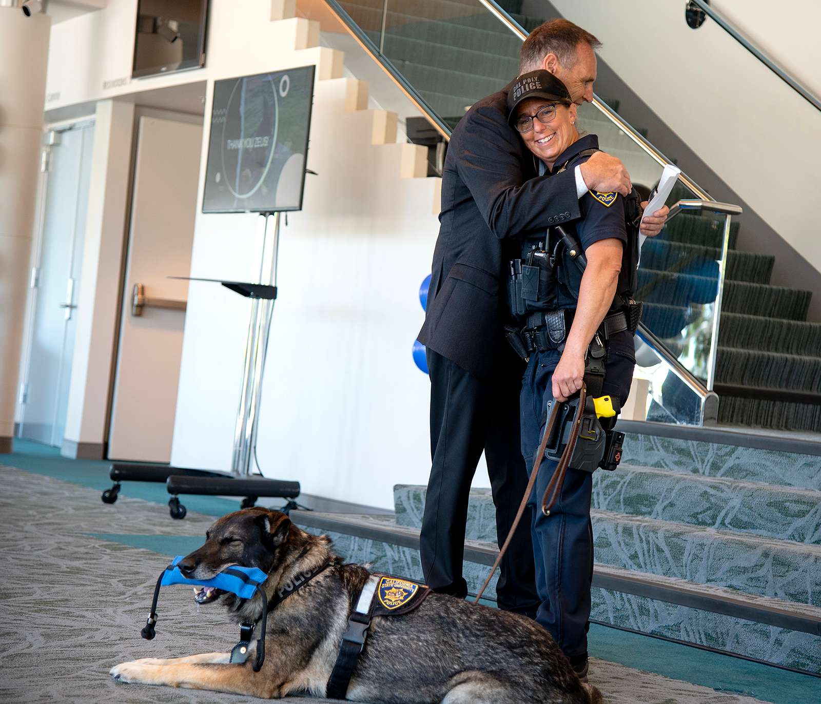 Cal Poly police officer hugs a colleague during a ceremony while a police K-9 wearing a vest lies on the floor chewing a toy.
