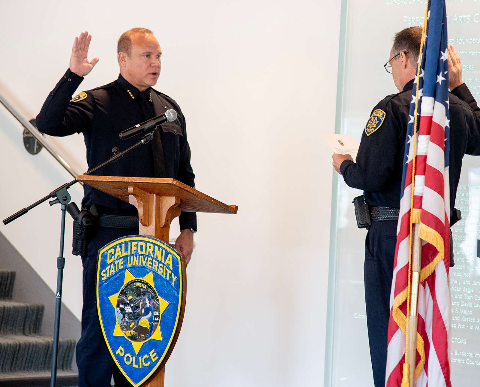 California State University police chief stands at a podium with one hand raised, taking the oath of office from another uniformed officer beside an American flag.