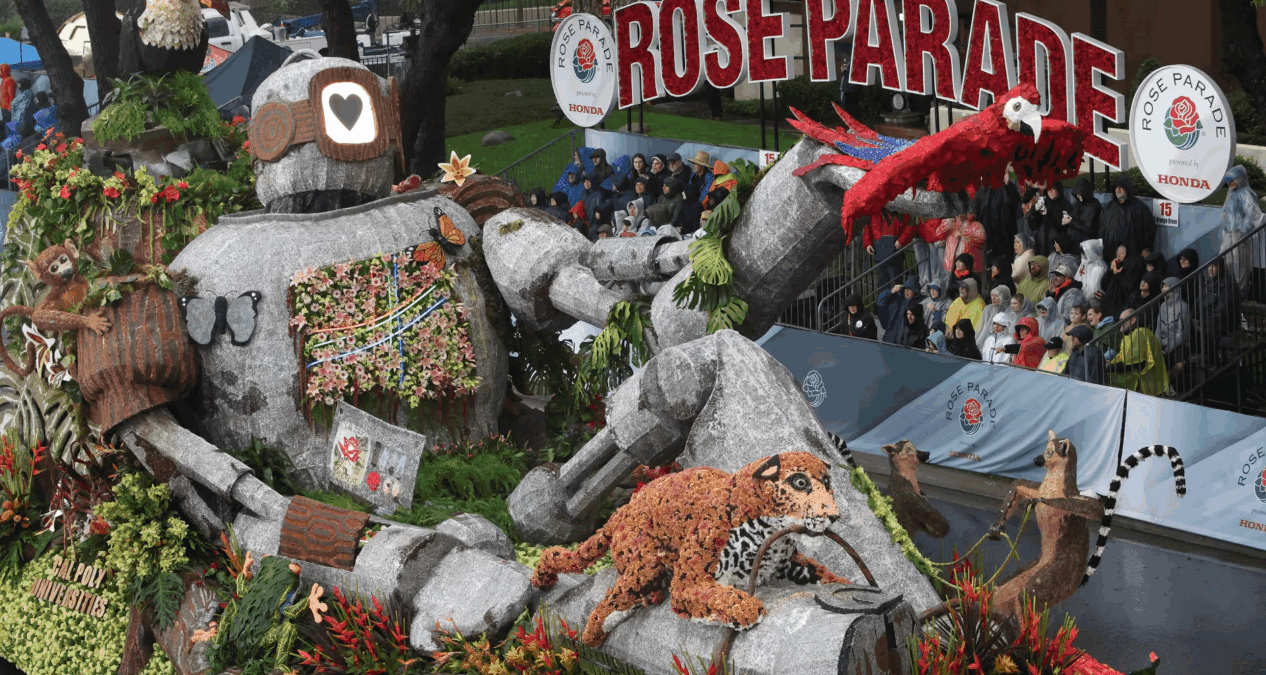 Floral float with large robot and various animals at the Rose Parade, surrounded by audience and Rose Parade signs.